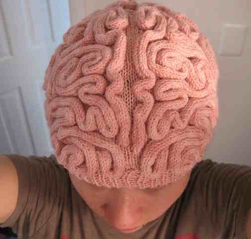 A close-up of a knitted hat shaped like a human brain, featuring a pink stockinette base with textured i-cord stitched on top to create anatomical squiggles and ridges.