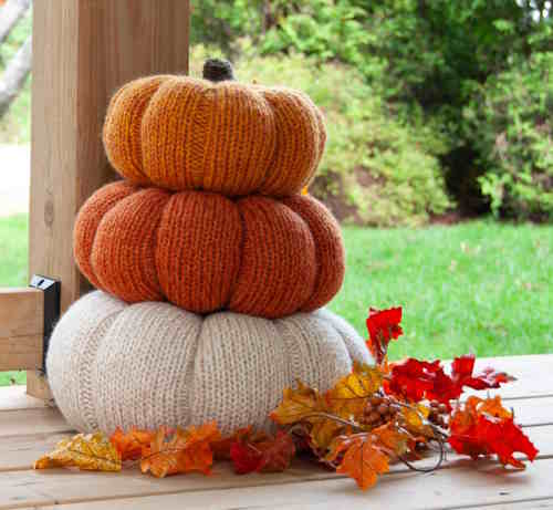 A stack of three large knitted pumpkins in varying shades of marled orange, featuring deep purl-stitch ribs and green knitted stems, displayed as outdoor fall porch decor.