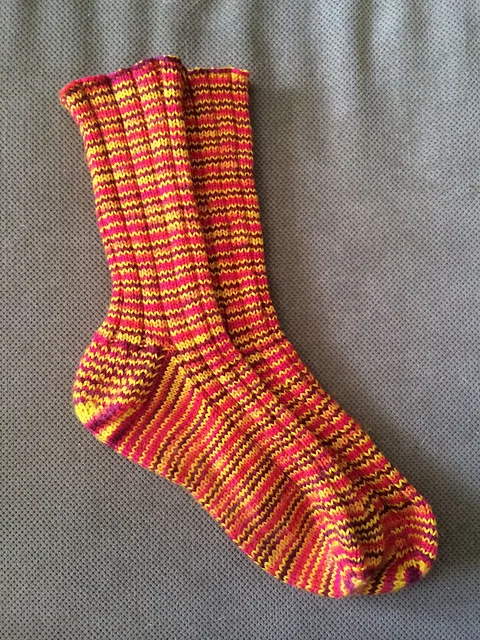 A pair of knitted socks featuring a continuous 2x2 ribbed texture from cuff to toe, worked in a variegated green and purple yarn, resting flat to display a traditional French heel turn.