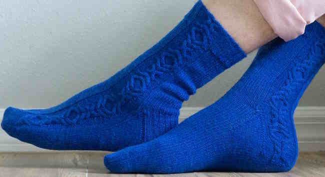 A close-up of a knitted sock in a rich jewel-toned blue yarn, featuring a thick chunky cable running down the leg and a smaller mirrored cable traveling down the instep.