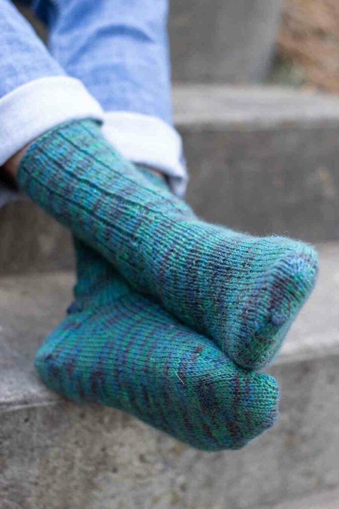 A pair of textured, variegated green socks featuring a stretchy K3, P1 ribbed cuff, knit with a chunky superwash merino wool yarn.