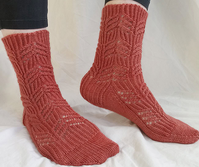 An orange-red knitted sock displaying a diagonal twisted stitch rib pattern interspersed with delicate lace eyelets, designed to mimic cascading water, with a matching blue variegated sock resting beside it.